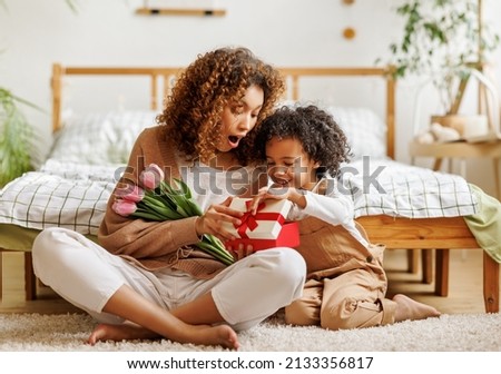 Cheerful african american family mom opens a gift box with her son and holding bouquet  of flowers while resting on floor by bed during holiday celebration mothers day at home Royalty-Free Stock Photo #2133356817
