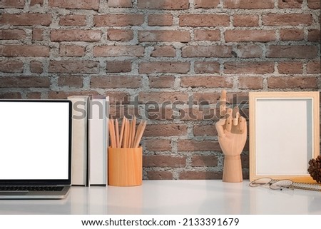 Computer laptop, picture frame, coffee cup and stationery on white table with brick wall.