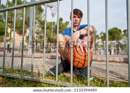young athlete with a basketball in his hand sitting on the basketball court behind the metal fence