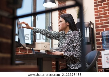 Young asian credit officer in financial company modern workspace interior sticking relevant information on work computer screen. Company office confident accountant putting sticky notes on PC monitor.