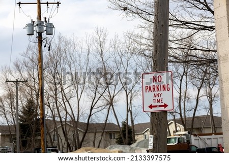 View of wooden utility pole and housing with No Parking Sign posted. Other poles with transformers attached. Bare tree branches with dump truck and snow pile. Blue sky with fluffy clouds. 