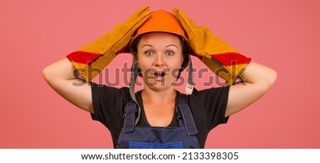 shocked woman in protective overalls, gloves and a helmet clutched her head with her hands on a pink background