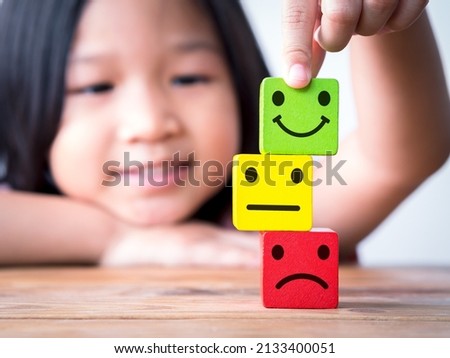 Little Asia child girl putting happy smiley face on top of neutral and sad or unhappy face icon on wood cube. Concept of happy children, kid, family, childrens day. Royalty-Free Stock Photo #2133400051