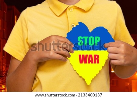 Inscription STOP WAR.Boy holding flag of Ukraine with the heart shape symbol.the city background is blurry.Selective focus.