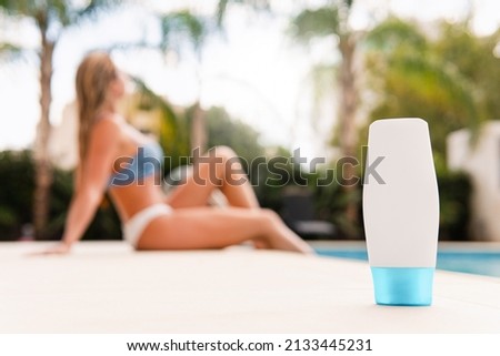White plastic bottle of skin care product with a young woman in bikini resting near the pool on background.