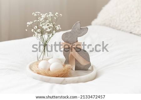 Marble tray, nest with white eggs, decorative bunny figure and vase with gypsophila flowers. White bedroom with Easter decoration. 