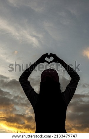 silhouette of young woman with her hands raised above her head makes a heart-shaped symbol against a beautiful twilight sky background. Heart symbol concept means love, friendship and kindness.