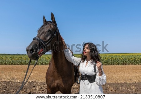Beautiful confident woman in white shirt is grooming brown horse. They are standing in agricultural field. Sunny day. Rural scene.