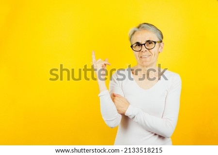 Old woman pointing signaling something with his finger, blank space for text or images posing isolated over yellow color wall background. Daily expressions with copy space in studio.