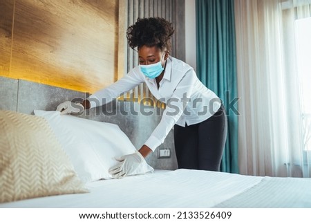 Chambermaid wearing protective face mask and gloves as precaution against coronavirus while working at a hotel. Beautiful maid with medical mask changing bed linen on the bed in a hotel room.