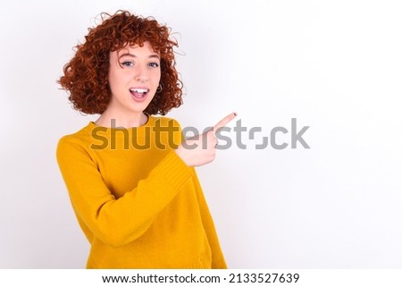 young redhead girl wearing yellow sweater over white background pointing away and smiling to you. Look over there!