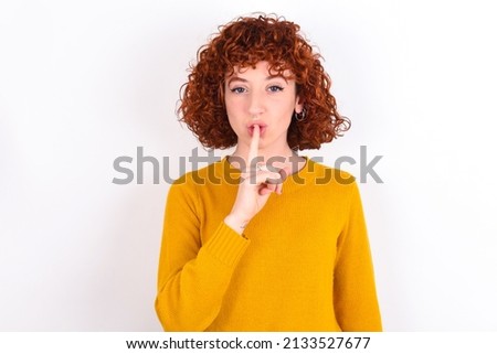 young redhead girl wearing yellow sweater over white background makes hush gesture, asks be quiet. Don't tell my secret or not speak too loud, please!