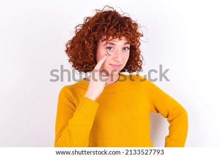 young redhead girl wearing yellow sweater over white background , looking, observing, keeping an eye on an object in front, or watching out for something. Royalty-Free Stock Photo #2133527793