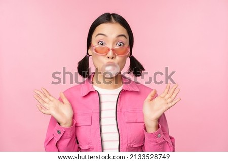 Stylish asian girl blowing bubblegum bubble, chewing gum, wearing sunglasses, posing against pink background Royalty-Free Stock Photo #2133528749