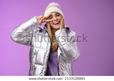 Positive lucky cute blond girl having fun winter vacation in silver glittering jacket hat posing happily smiling white teeth enjoying trip show peace victory sign near eye, standing purple background