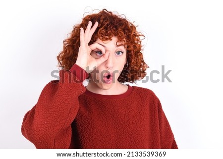 young redhead girl wearing red sweater over white background doing ok gesture shocked with surprised face, eye looking through fingers. Unbelieving expression.
