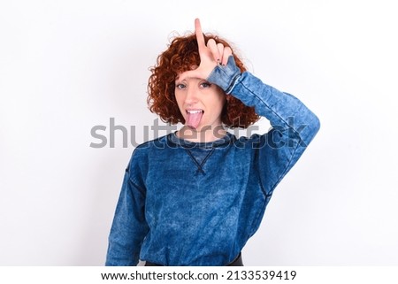 young redhead girl wearing blue sweater over white background gestures with finger on forehead makes loser gesture makes fun of people shows tongue