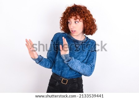 Displeased young redhead girl wearing blue sweater over white background keeps hands towards empty space and asks not come closer sees something unpleasant