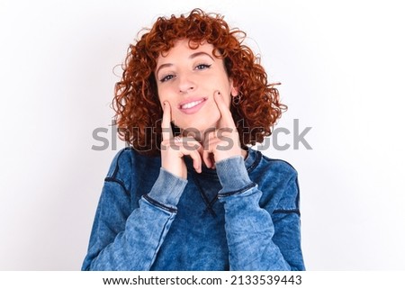 Happy young redhead girl wearing blue sweater over white background with toothy smile, keeps index fingers near mouth, fingers pointing and forcing cheerful smile