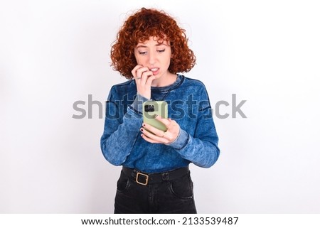 Portrait of pretty frightened young redhead girl wearing blue sweater over white background chatting biting nails after reading some scary news on her smartphone.