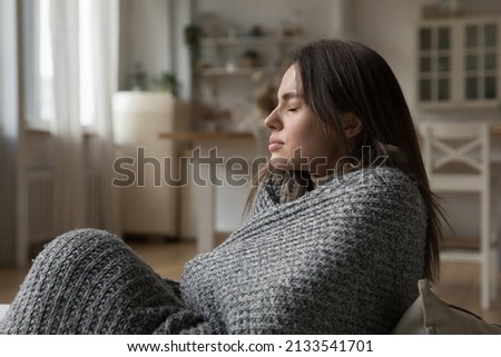 Young woman covered in warm knitted plaid sits on sofa at home suffers from low temperature in living room, shivers, having flu symptoms feeling unwell. Heating problem, catch cold, discomfort concept Royalty-Free Stock Photo #2133541701