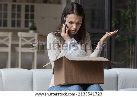 Dissatisfied angry woman, cheated client sit on sofa check received box, damaged or broken goods in parcel, talks to customers support, express complaints, looks annoyed. Bad delivery services concept Royalty-Free Stock Photo #2133541723