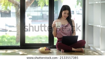 Attractive young woman holding coffee cup and using digital tablet while sitting on wooden floor in living room.