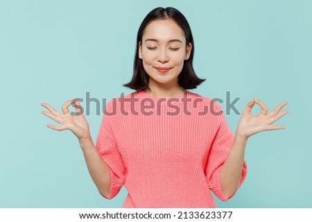 Young spiritual happy woman of Asian ethnicity 20s wearing pink sweater hold spreading hands in yoga om aum gesture relax meditate try to calm down isolated on pastel plain light blue color background