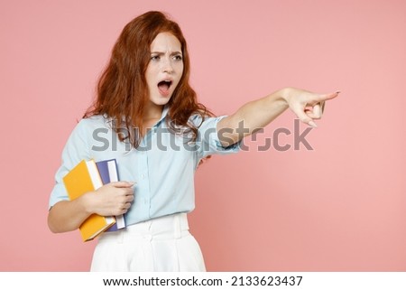 Young angry indignant redhead student woman in blue shirt hold book point index finger aside command isolated on pastel pink background studio portrait Education high school university college concept