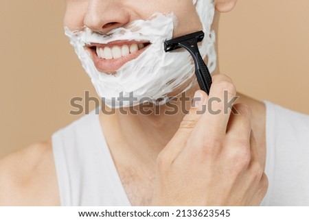 Cropped smiling young man 20s perfect skin in undershirt face covered with foam shaving with blade isolated on pastel pastel beige background studio Skin care healthcare cosmetic procedures concept Royalty-Free Stock Photo #2133623545