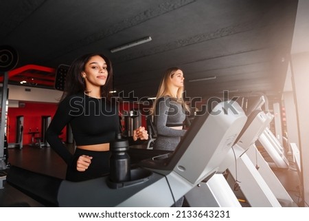 Two young beautiful girls with slim sport figure running on treadmill in modern gym. Healthy and sporty lifestyle concept. Side view.