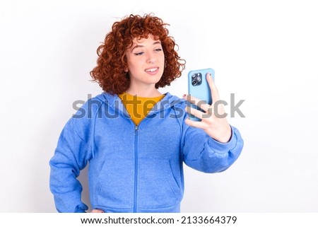 Isolated shot of pleased cheerful young redhead girl wearing blue jacket over white background , makes selfie with mobile phone. People, technology and leisure concept