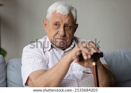 Lonely tired depressed senior old man sitting on sofa home, holding walking cane, looking away with sad unhappy face, suffering from arthritis disability, feeling pain, frustration. Elderly healthcare Royalty-Free Stock Photo #2133684203