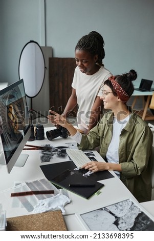 Vertical portrait of two smiling female photographers discussing images on computer screen while working on editing in studio
