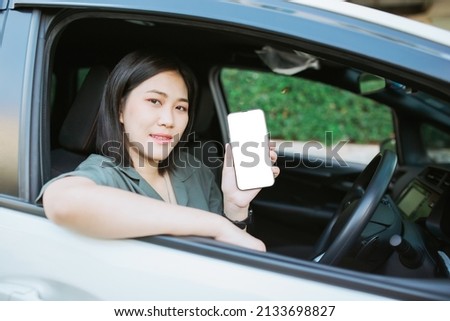 Young Asian beautiful woman sitting in her car. Young Asian beautiful woman wears a green shirt and smiling. Young Asian beautiful woman showing a blank screen smartphone.Concept insurance