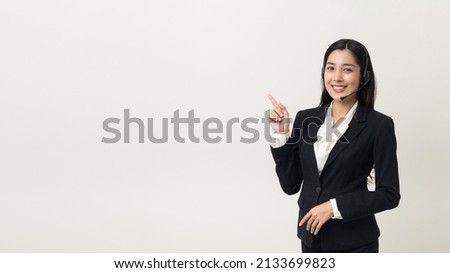 Happy young asian business woman call centre pointing finger to blank space. Welcome female operator put on smalltalk headphone standing on isolated white background. Royalty-Free Stock Photo #2133699823