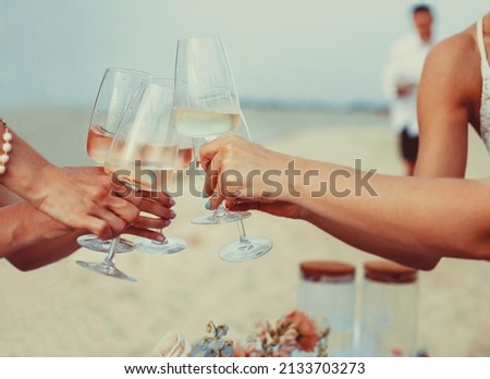 Happy female friends in summer dresses smiling and clinking glasses of wine while resting on beach together Royalty-Free Stock Photo #2133703273