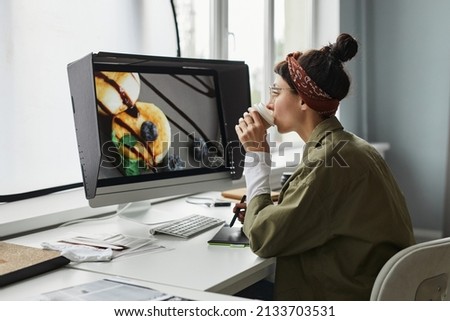 Side view portrait of young female photographer editing photos while using laptop in studio and drinking coffee