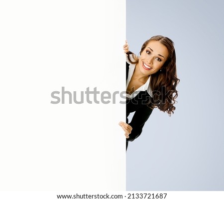 Happy smiling brunette young business woman in blue clothing, standing behind, peeping from blank banner or mock up signboard, showing copy space for text, over gray background.
