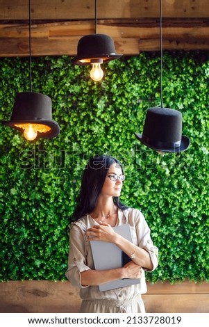 Portrait modern female woman using laptop and phone, communicates on internet with customer in city cafe. Cozy office workplace. Eco-friendly background wall of green grass