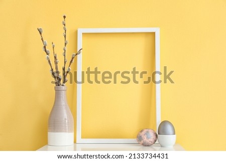Empty picture frame, vase with pussy willow branches and Easter eggs on color background