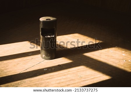 An old metal trash bin stands in the middle of a room of an abandoned building, the bin is stuffed with cigarette butts and ashes, the bright sun illuminates this unpretentious picture