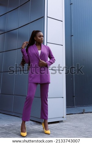 Outdoor full body length portrait of African girl with braids and make up wearing purple stylish pants suit and posing at urban minimalist grey background. Diversity fashion lifestyle concept.