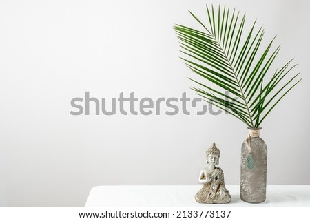 Modern minimal interior floral decoration with natural palm leaves in vintage glass vase and Buddha statue on white linen table cloth. Light wall as background with copy space