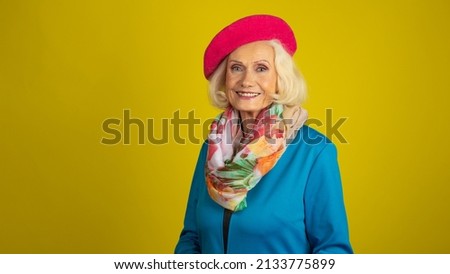 Adorable Elderly Woman Posing on a Yellow Back Ground Set while Wearing a Bright Blue Top and a Red Barrette and Giving a Big Smile.