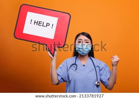 Young adult hospital nurse holding cardboard speech bubble sign while wearing stethoscope and virus protection facemask. Worried medical staff worker wearing medical instrument and asking for help. Royalty-Free Stock Photo #2133781927