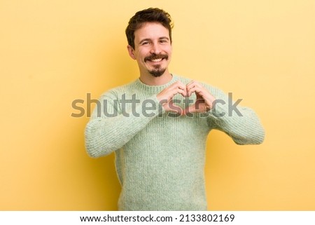 young hispanic man smiling and feeling happy, cute, romantic and in love, making heart shape with both hands