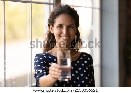 Portrait of smiling latin woman holding glass of cool mineral water, proposing drinking every day, keeping organism refreshed preventing dehydration, feeling satisfied with healthcare lifestyle. Royalty-Free Stock Photo #2133836521