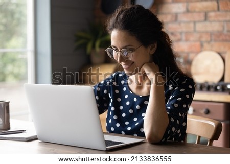 Happy young hispanic female freelance worker employee entrepreneur in eyeglasses looking at computer screen, working on creative online project distantly at home, reading email with pleasant news.