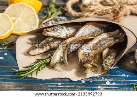 Sun-dried smelt in a paper package. Salted fish with marine decor. Lemon, fresh rosemary, sea rope. Blue nautical wooden background, close up Royalty-Free Stock Photo #2133892793